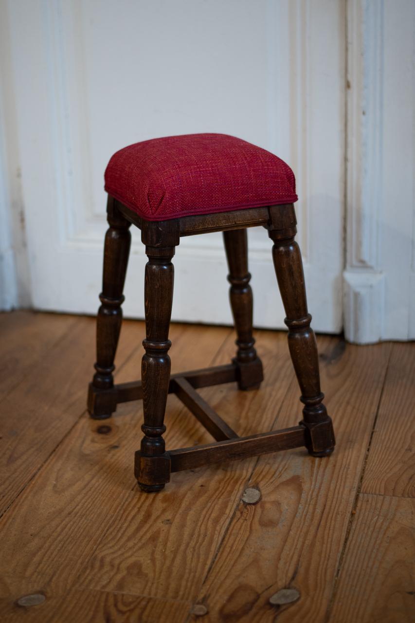 petit tabouret avec des pieds tournés recouvert de tissu 
            rouge. Finition : galon double corde rouge